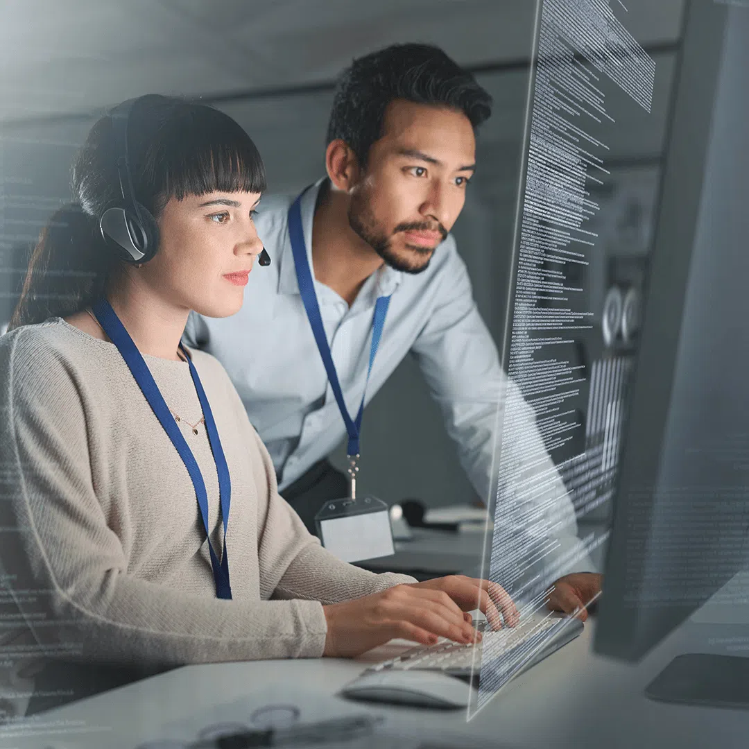 Two colleagues wearing lanyards work together at a computer, focusing on code displayed on the screen in a modern office setting.
