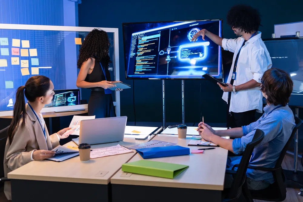 A group of four people in a modern office meeting room collaborate around a large digital display showing software code, cloud computing graphics, and technology diagrams. One person stands at the screen presenting with a tablet while three others sit at a conference table with laptops, notebooks, and office supplies. Sticky notes are visible on a glass wall in the background, suggesting a planning or development session. The room is lit with cool blue tones that emphasize the technology-focused environment.