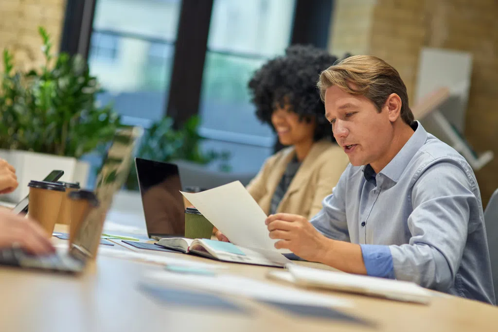 Person reviewing a document at a meeting table with laptops, notebooks, and coffee cups in a modern office setting.