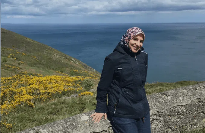 Nancy Hendy sat on a wall in front of a sea and fields landscape
