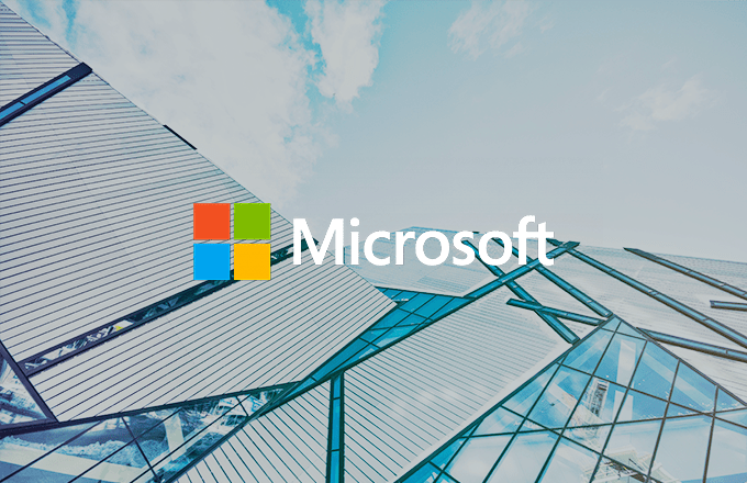 Microsoft logo overlaid on a modern glass office building with geometric panels, viewed from below against a light blue, partly cloudy sky.