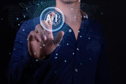 A person in a dark blue shirt is interacting with a futuristic holographic interface. They are pressing a virtual button that displays an icon of a brain within a circle, with the word AI in it. The background features digital binary code and circuit lines, suggesting advanced technology or computing concepts. There are glowing dots connected by lines that create a network pattern around the person’s hand and the holographic display, enhancing the high-tech atmosphere of the image.
