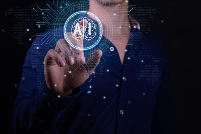 A person in a dark blue shirt is interacting with a futuristic holographic interface. They are pressing a virtual button that displays an icon of a brain within a circle, with the word AI in it. The background features digital binary code and circuit lines, suggesting advanced technology or computing concepts. There are glowing dots connected by lines that create a network pattern around the person’s hand and the holographic display, enhancing the high-tech atmosphere of the image.
