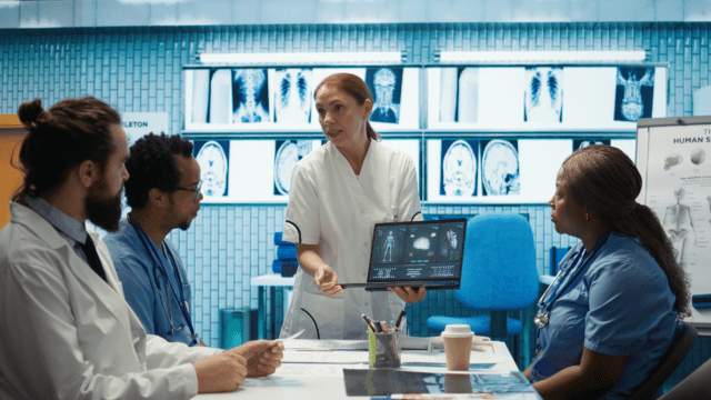 A diverse medical team of healthcare professionals around a table with the front speaker standing, holding a tablet with AI generated imagery of anatomy.
