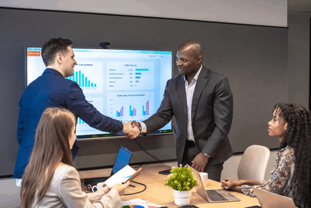 Four professionals in a conference room during a business meeting; two men shake hands in front of a screen while two women work on laptops at the table.