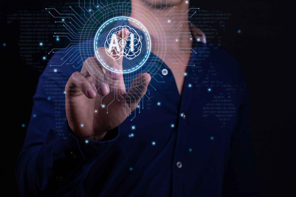 A person in a dark blue shirt is interacting with a futuristic holographic interface. They are pressing a virtual button that displays an icon of a brain within a circle, with the word AI in it. The background features digital binary code and circuit lines, suggesting advanced technology or computing concepts. There are glowing dots connected by lines that create a network pattern around the person’s hand and the holographic display, enhancing the high-tech atmosphere of the image.