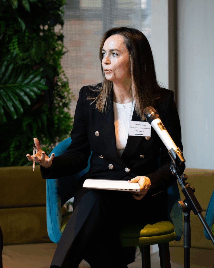 A speaker seated on a chair, holding a pen and notebook, speaking into a microphone during a Women in Tech Leadership event. A green plant wall is visible in the background.