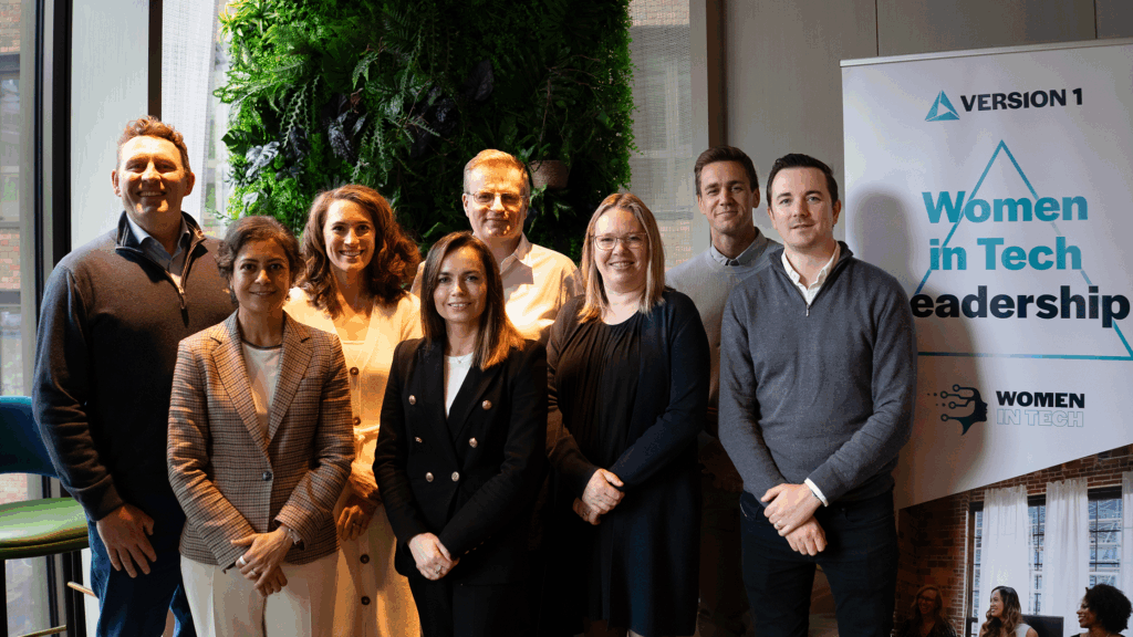 Group of eight people standing together indoors in front of a green plant wall and a banner that reads ‘Version 1 Women in Tech Leadership’.