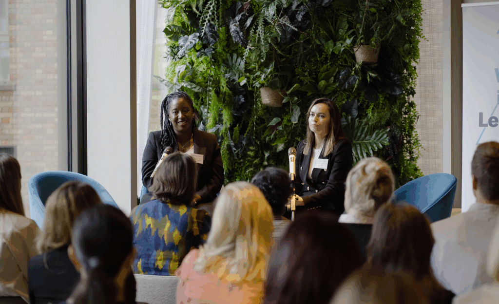Two speakers seated on stage in front of a green plant wall, addressing an audience at a Women in Tech Leadership event. A banner with the event name is partially visible on the right.
