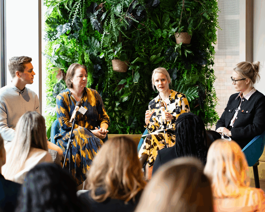 Four speakers seated in front of a lush green plant wall, engaged in a panel discussion at a Women in Tech Leadership event. Audience members are visible in the foreground.