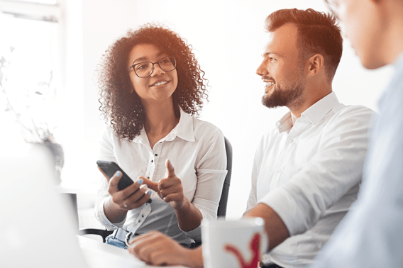 wo people sitting together at a desk in a bright office, discussing something while one holds a smartphone and the other has a coffee mug. A third person is partially visible on the right side of the image.