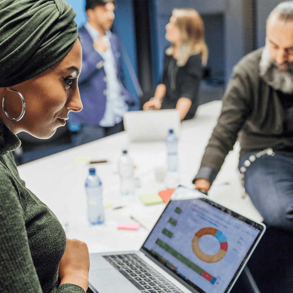“Woman wearing a headscarf reviews analytics on a laptop during a team meeting, with colleagues discussing in the background.