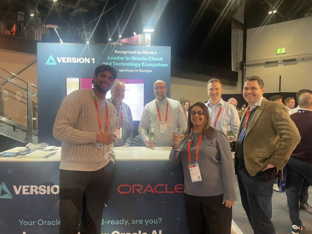 A group of several individuals stands in front of a booth at Oracle AI World Tour London 2026. The booth features prominent signage for “Version 1” and “Oracle,” along with text highlighting leadership in Oracle Cloud and technology ecosystem services in Europe. The individuals wear event lanyards and hold drinks while posing together. Behind them, additional attendees and exhibition stands are visible in a well‑lit, modern venue.