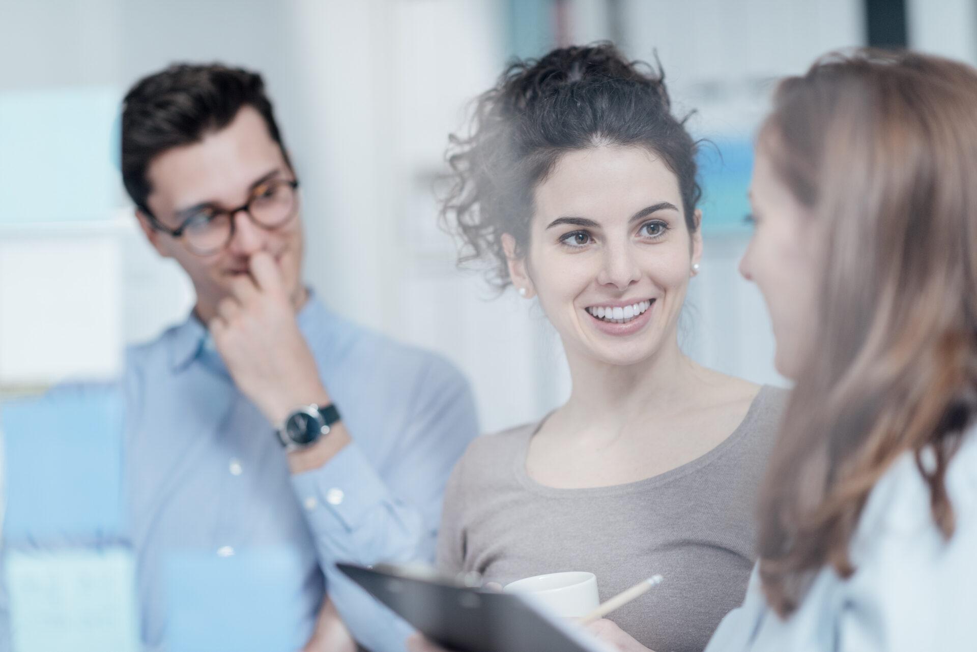 Three colleagues in a modern office discussing work, with a woman holding a tablet and smiling during a team conversation.