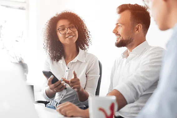 wo people sitting together at a desk in a bright office, discussing something while one holds a smartphone and the other has a coffee mug. A third person is partially visible on the right side of the image.