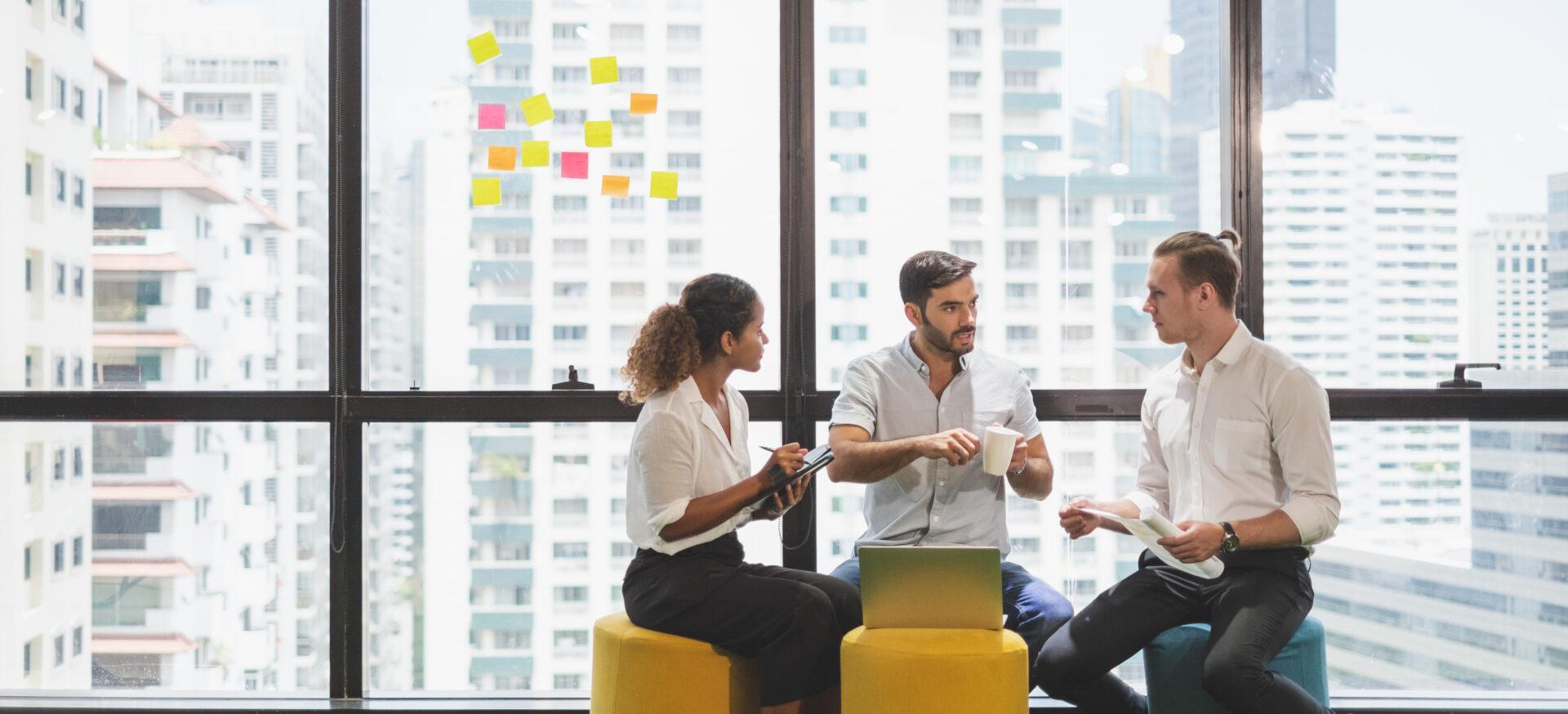 A group of three people sit on colorful stools in a modern office space, talking and holding work materials. A laptop is open on one of the stools, and sticky notes cover the window behind them with tall city buildings visible outside.