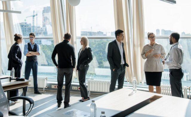 Group of professionally dressed colleagues networking and chatting in a modern office with large floor-to-ceiling windows overlooking a city skyline.