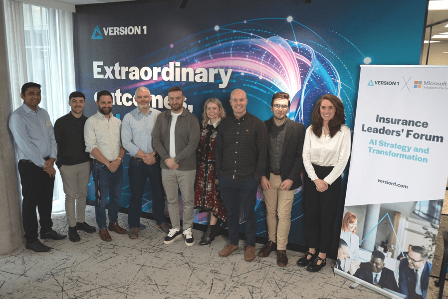 A group of nine professionals standing together at a corporate event, posing in front of a large “Version 1” branded backdrop with a colourful digital swirl design and the words “Extraordinary outcomes.” To the right, a banner promotes an “Insurance Leaders’ Forum: AI Strategy and Transformation” in partnership with Microsoft. The group is dressed in smart casual business attire and smiling at the camera.