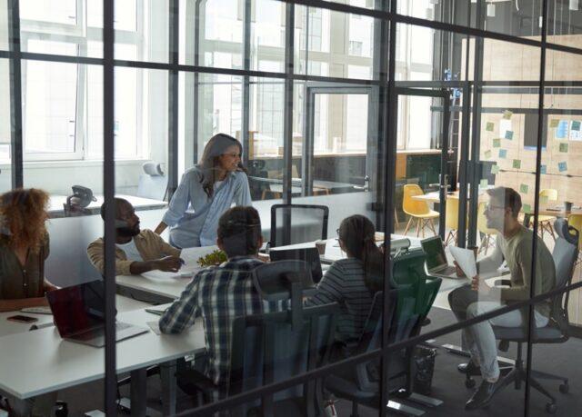 Team of colleagues collaborating in a modern glass-walled office meeting room, seated around a table with laptops and documents while a woman stands and speaks, and another person works in the background near a wall covered with sticky notes.