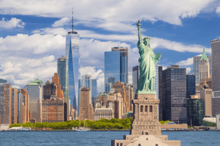 View of the Statue of Liberty in the foreground with the Manhattan skyline and One World Trade Center in the background under a partly cloudy sky.
