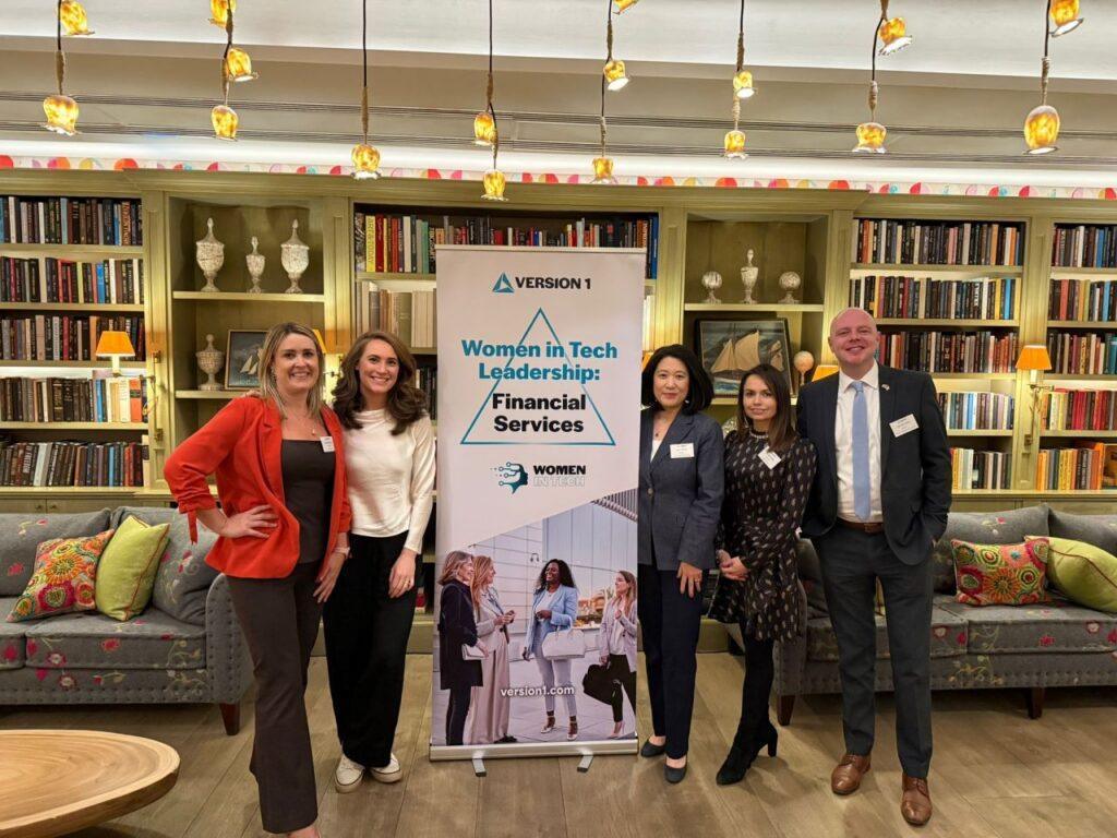 Group of five professionals standing in a stylish lounge with bookshelves and warm lighting, posing beside a banner that reads “Women in Tech Leadership: Financial Services” by Version 1.