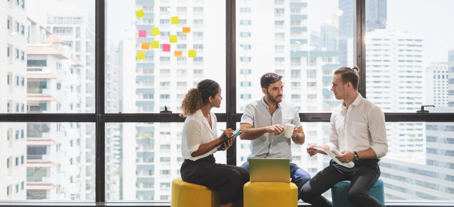 A small group of three colleagues sit together in a bright office lounge area in front of large windows overlooking tall city buildings. They are talking and sharing ideas while using a laptop, with colourful sticky notes arranged on the window behind them.