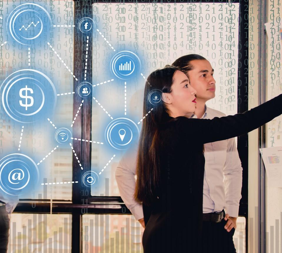 A group of people stand in front of large windows with a digital overlay of connected icons symbolizing communication, finance, data, and social networks. Blue circular icons—such as a dollar sign, an email symbol, a location marker, and social media symbols—are linked by dotted lines across a background of binary code. One person in the foreground gestures toward the digital display while others stand nearby, suggesting a discussion or presentation related to technology or data networks.