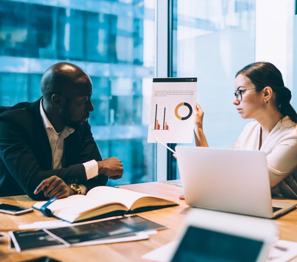 A person seated at a conference table listens while another person holds up a printed page displaying bar charts and a circular chart. The table is covered with a laptop, open notebooks, and documents. Large windows in the background reveal the exterior of a modern office building, creating a professional meeting setting focused on data or performance review.Provide your feedback on BizChat