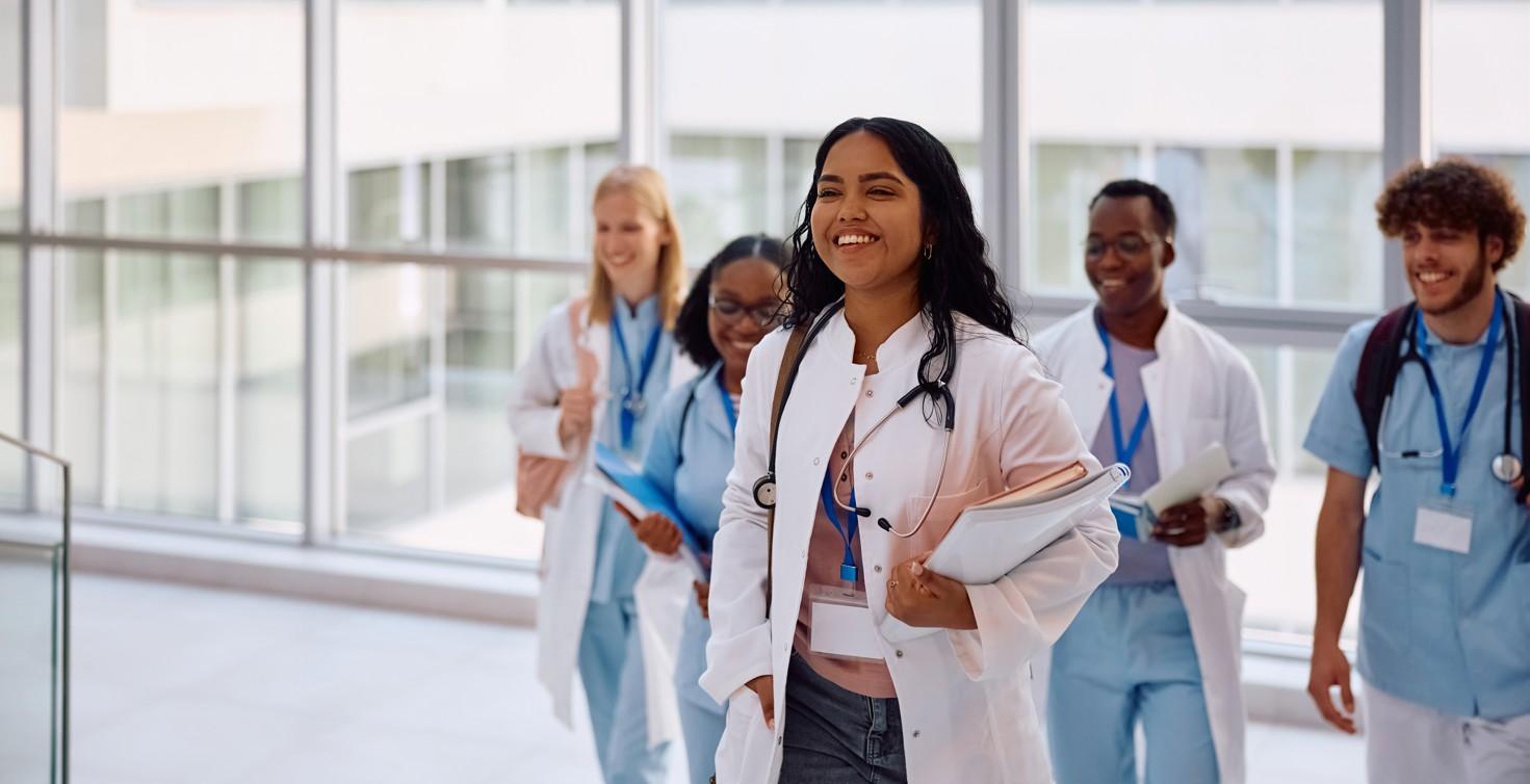 A group of individuals wearing medical coats and scrubs walk together through a bright hallway with large windows. One person in front carries folders and a bag, while others behind hold notebooks or wear backpacks. The setting appears to be a modern healthcare or academic facility.