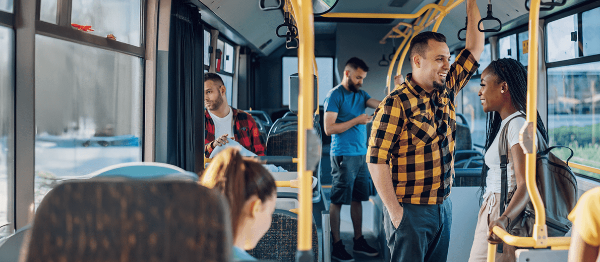 Passengers ride on a city bus, with some seated and others standing and holding onto overhead rails. Two people near the front are engaged in conversation, while others look at their phones or out the windows.