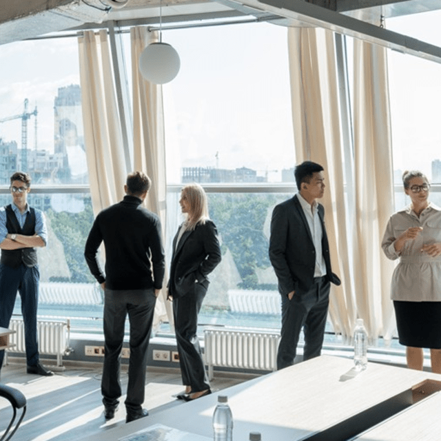 A group of people in business attire stand and talk in a modern office space with large floor‑to‑ceiling windows. Sunlight fills the room, and an urban skyline with trees and buildings is visible outside. Office desks, chairs, and bottles of water are in the foreground.