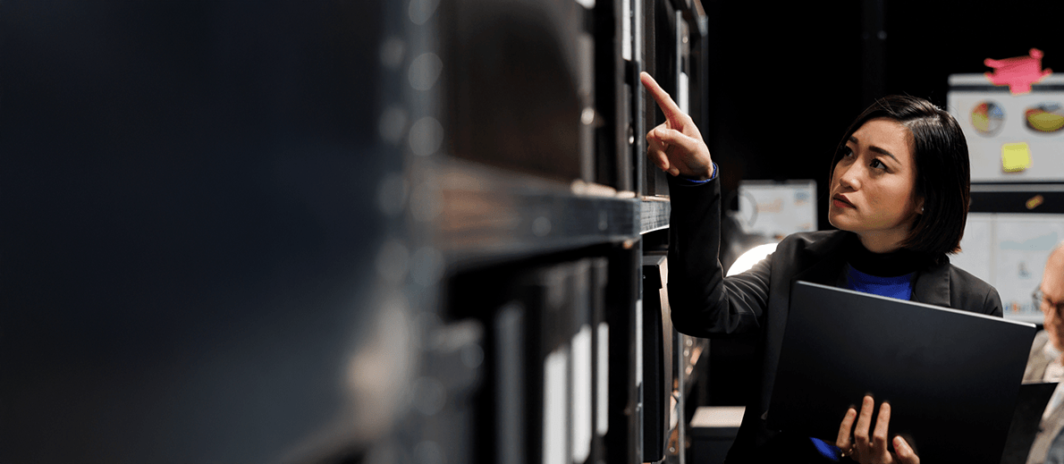 A person stands in front of a shelf full of binders, holding a folder and pointing toward one of the items on the upper shelf, as if locating or retrieving a document.