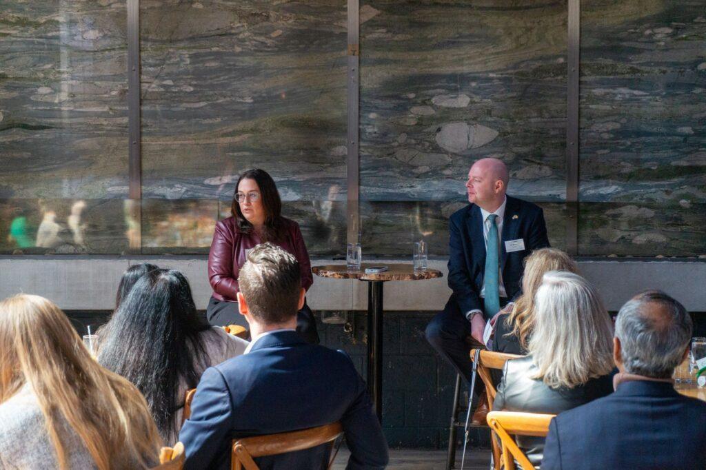 Two speakers seated at a small table address an audience at Version1's Women in Tech Leadership NJ: Pharmaceuticals, MedTech & Life Sciences event, with attendees listening in an indoor setting.