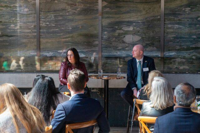 Two speakers seated at a small table address an audience at Version1's Women in Tech Leadership NJ: Pharmaceuticals, MedTech & Life Sciences event, with attendees listening in an indoor setting.