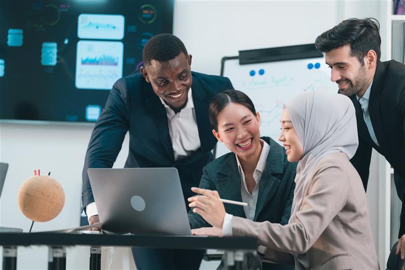 A group of four professionals collaborates around a laptop in a modern office setting. One person gestures with a pen while others lean in to review information on the screen. Behind them, charts and graphs are displayed on a monitor and a whiteboard, suggesting a data-focused discussion or meeting.