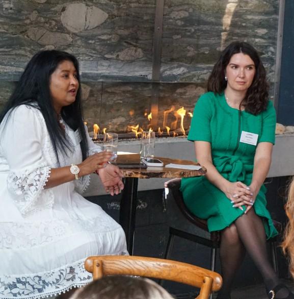 Two adults sit on stools at a small round table indoors, speaking during a discussion. One wears a white outfit and gestures while holding a glass of water; the other wears a green dress and sits with hands folded. A modern stone wall with a narrow, horizontal fireplace burning behind them forms the background, with glasses and a phone on the table in between. Women in Tech Leadership