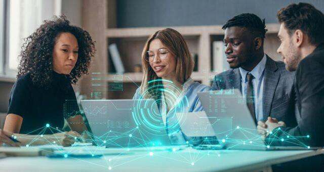 A group of four people sit around a table in a modern office environment, collaborating while using laptops and documents. A digital-looking overlay with graphs, data visualizations, and interface elements is superimposed in the foreground, giving the scene a futuristic or technology‑focused feel.