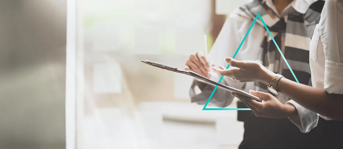 Two people collaborate while reviewing documents on a clipboard, one pointing with a pen as they discuss details. A geometric triangle graphic overlays the scene, adding a modern design element.