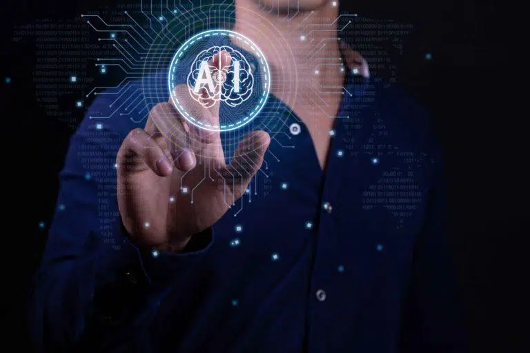 A close-up, futuristic-themed image shows a person wearing a dark shirt interacting with a transparent digital interface. The person’s index finger is extended toward the center of the image, as if tapping or selecting a virtual button. In front of the fingertip, a glowing circular icon displays the letters “AI” over a stylized brain graphic split into two hemispheres. Surrounding the central icon are circuit-like lines and patterns, suggesting a digital network or data flow. The background is dark blue with scattered glowing points of light and faint binary-style text, giving a high-tech, artificial intelligence and data-processing atmosphere.