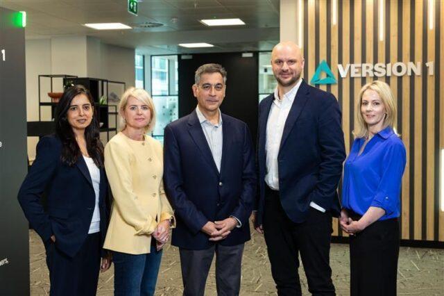 Group photograph of six executive team members standing in a modern office reception area, with the Version 1 logo visible on the wall behind them.