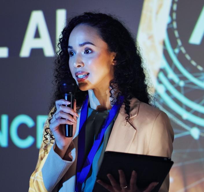 Professional speaking at a conference, holding a microphone and tablet, presenting on AI and cybersecurity topics in a modern tech setting.