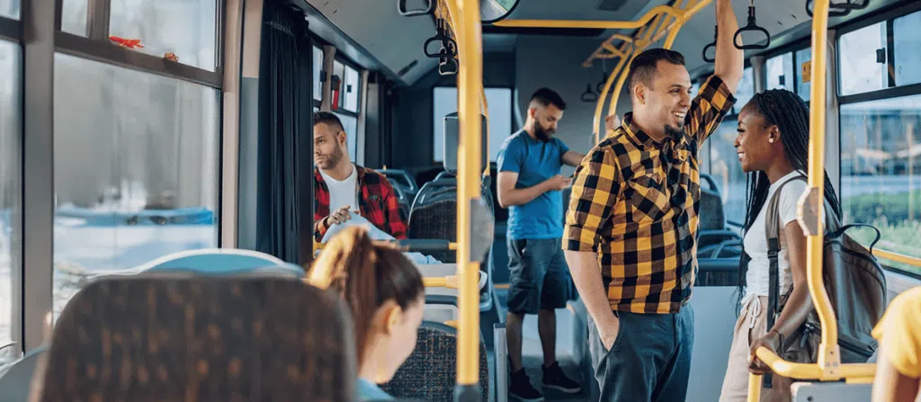 A photograph taken inside a public bus during daylight, showing several passengers and the interior layout. In the foreground, there are yellow vertical and horizontal handrails typical of public transport seating areas. Near the center-right, one passenger stands facing another passenger. The standing passenger is holding onto an overhead rail with one hand while wearing a yellow-and-black checkered long-sleeve shirt and dark trousers. The other nearby passenger, standing and facing toward the first, has long braided hair and is wearing a light-colored top with a backpack over one shoulder. In the background, additional passengers are seated and standing. On the left side, one seated passenger is wearing a red plaid shirt and looking downward at an object held in both hands. Further back in the aisle, another standing passenger is wearing a blue T-shirt and also looking down at a handheld device. The bus interior includes rows of grey seats, overhead hand grips, and large windows along the side allowing natural light into the space. Through the windows, an urban outdoor setting is faintly visible but not clearly detailed. The scene shows a typical moment of public transit use, with passengers standing, sitting, and holding onto support bars while the bus is in motion.