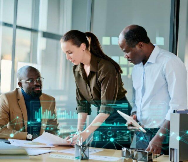 Three business professionals in an office reviewing documents together, with one person pointing at papers while others look on, overlaid with digital data visuals suggesting analytics or AI-driven insights.