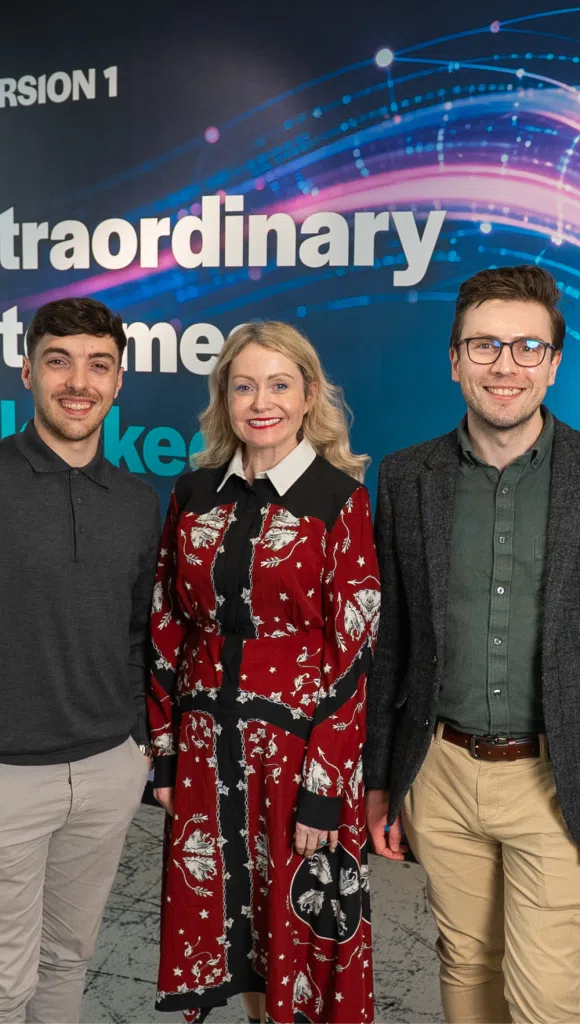 Three professionally dressed adults stand side by side, smiling at the camera in front of a blue event backdrop with abstract digital wave graphics and partial text.