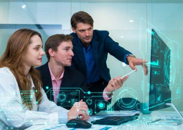 Three individuals in a modern office collaborate at a desk while viewing a computer display with futuristic, holographic-style data visualisations. One individual stands and points toward the screen as the other two sit and work together. Image used for an Oracle AI webinar page.