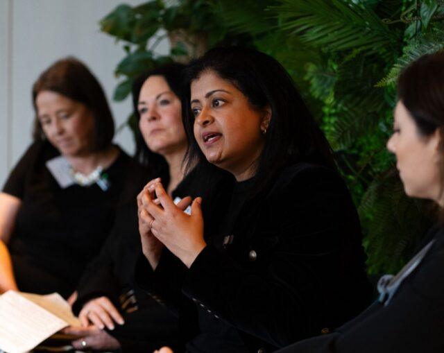 A speaker addressing attendees during Version 1&rsquo;s Women in Tech Leadership event, standing in front of a green plant wall and engaging the audience during a fireside-style discussion.