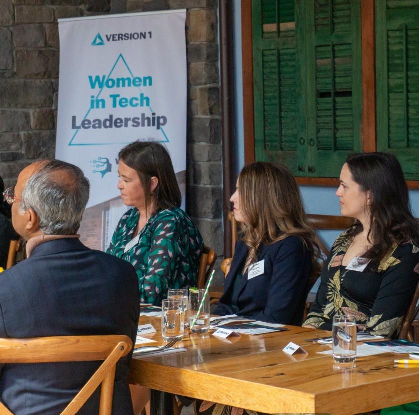 Several adults are seated around a wooden table at an indoor event, facing the same direction. Glasses of water, paper name cards, and printed materials are on the table. Behind them, a standing banner reads “Women in Tech Leadership,” and the background includes exposed brick walls and green wooden shutters. Women in Tech Leadership