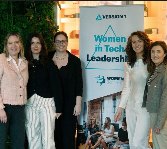 A group of five attendees standing side by side at a professional event, posing in front of a banner that reads “Version 1 Women in Tech Leadership.” The setting includes indoor greenery and warm lighting, with the banner and event space visible in the background.