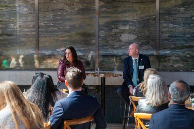 Two speakers seated at a small table address an audience at Version1's Women in Tech Leadership NJ: Pharmaceuticals, MedTech & Life Sciences event, with attendees listening in an indoor setting.