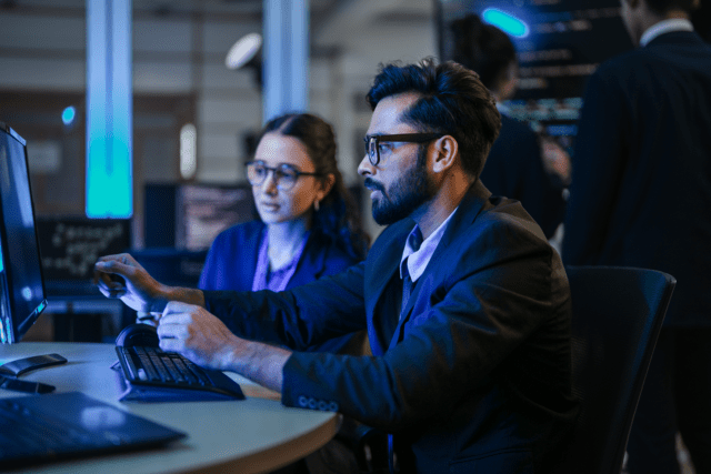 A professional cyber security team, including a man and a woman in formal attire, works intently in a high-tech, dimly lit command center. The man in the foreground, wearing glasses and a suit, focuses on a glowing monitor while his colleague observes, highlighting a collaborative and high-stakes environment.
